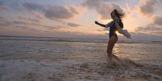 快乐的女人穿着泳衣和衬衫在日落时走在海滩上。海浪拍打着女性的脚。年轻美丽的女孩享受生活，在海边玩得开心。暑假。慢动作