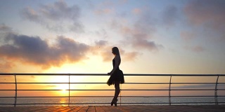 一个穿着黑色芭蕾舞裙的芭蕾舞女演员在日出或日落时在海洋或海滩的堤岸上跳着芭蕾舞。剪影的年轻美丽的金发女人长头发练习拉伸和练习。慢动作