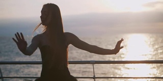 一个穿着黑色芭蕾舞裙的芭蕾舞女演员在日出或日落时在海洋或海滩的堤岸上跳着芭蕾舞。年轻美丽的金发女子与长头发练习伸展和练习。慢动作