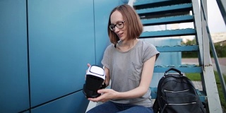 一个戴眼镜的小女孩手里拿着一副蓝色背景的虚拟现实眼镜。带着vr眼镜坐在楼梯上的微笑女人。