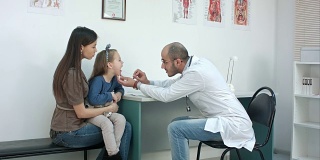男儿科医生检查咽喉的小女孩坐在她的母亲