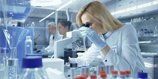 女性研究科学家在显微镜下观察生物样本。她和她的同事们在一家大型现代化实验室/医疗中心工作。