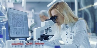 女研究科学家在显微镜下观察生物样本，将结果输入计算机。她和她的同事们在一家大型现代化实验室/医疗中心工作。