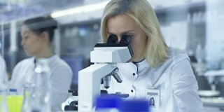 在显微镜下观察样品的女研究科学家们把滑片递给她的同事。他们在一个大型研究中心/实验室工作。
