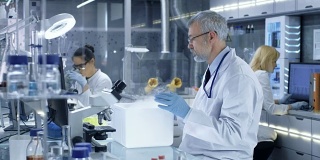高级医学研究科学家打开冰箱，拿出装有样品的培养皿并检查它。他在一个繁忙的现代化实验室中心工作。