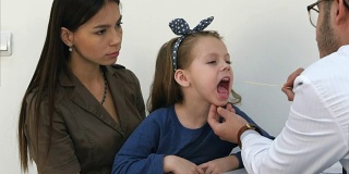 男儿科医生检查咽喉的小女孩坐在她的妈妈