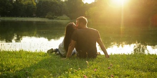 一对情侣在美丽的日落中接吻，坐在城市公园的湖边。外面快乐的夫妇的后视图