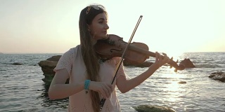年轻美丽的白人女孩穿着牛仔裤在海滩上拉小提琴靠近美丽的大海或海洋，慢镜头