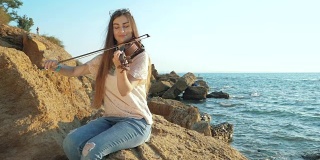 年轻漂亮的白人女孩穿着牛仔裤在美丽的海边拉小提琴，女人坐在大岩石上。慢动作