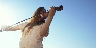 年轻美丽的白人女孩穿着牛仔裤拉着小提琴停留在蓝色的大海背景，慢镜头