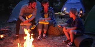 带着帐篷旅行，在森林里野餐，晚上一家人围着火堆准备黄色的玉米，妈妈、儿子和爸爸在这期间