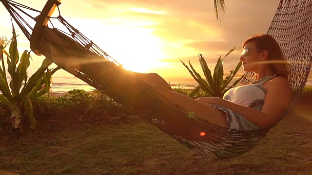 慢动作照明弹:黑发女子躺在摇摆的吊床上，在金色的日落海滩上