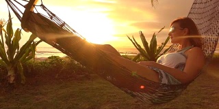 慢动作照明弹:黑发女子躺在摇摆的吊床上，在金色的日落海滩上