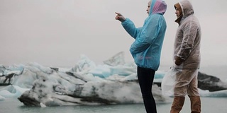 年轻的旅行夫妇穿着雨衣在冰岛的冰泻湖欣赏风景。游客在寒冷的日子里欣赏大自然