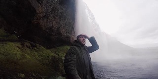 一个快乐的年轻人拿着自拍杆在大雾天用gopro相机拍摄强大的Seljalandsfoss瀑布