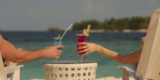 年轻的家庭夫妇日光浴和喝彩色鸡尾酒在热带海滩。情侣在蜜月异国度假躺在海边的躺椅上的阳光度假