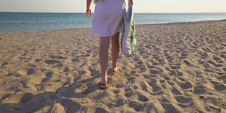 年轻的女孩穿着白色的夏装走过沙滩走向大海