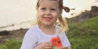 可爱的小女孩坐在池塘边的绿草上吃着红西瓜