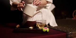美丽的黑发在浴衣拿茶壶和贫穷的茶在玻璃杯喝草药茶在温泉慢动作。茶道。Slowmotion拍摄