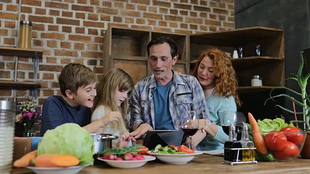 快乐家庭在厨房做饭时使用数字平板触摸屏父母和两个孩子在家交谈