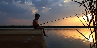 日落时分，小男孩坐在木码头边，在湖里钓鱼。