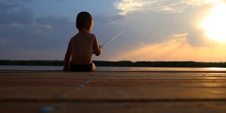 后视图的小男孩坐在一个木制码头的边缘和钓鱼的湖在日落。