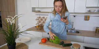 白天，迷人的女人站在时尚的厨房中央，手里拿着手机做饭，用蔬菜作图