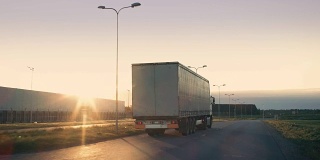 一辆带拖车的半卡车在高速公路上行驶的后续镜头。一辆白色卡车在阳光照耀下的空旷道路上穿过工业仓库。