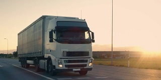 一辆带拖车的半卡车在高速公路上行驶的后续镜头。清晨，白色卡车驶过工业仓库。