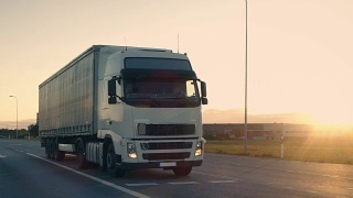 一辆带拖车的半卡车在高速公路上行驶的后续镜头。清晨，白色卡车驶过工业仓库。视频素材模板下载