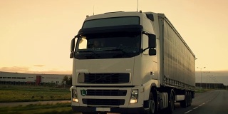前视摄像头跟踪半卡车与货运拖车在高速公路上行驶。他在夕阳的背景下快速穿过工业仓库。