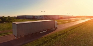在高速公路上移动的白色半挂车的鸟瞰图。在背景中可以看到仓库和工业装载建筑。日落。