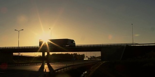 侧视图的白色半挂车与货物拖车驾驶过一座桥。美丽的风景，车辆行驶在天桥上。天黑了，太阳要下山了。