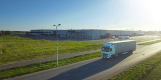 空中跟踪拍摄的白色卡车与半挂车连接移动通过工业仓库，农村地区。日落。