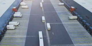 航拍一辆挂着半挂车的卡车离开工业仓库/仓库大楼/装卸区，那里有许多卡车在装卸货物。