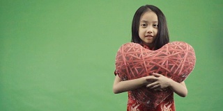 亚洲小女孩抱着孤立的心枕在绿色背景上，慢动作拍摄