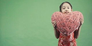 亚洲小女孩抱着孤立的心枕在绿色背景上，慢动作拍摄