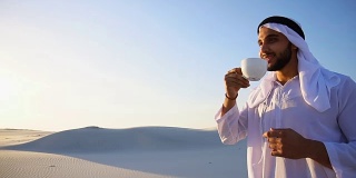 年轻英俊的阿拉伯酋长的肖像，他喝着咖啡，站在沙漠中，对着蓝天