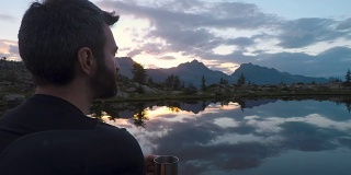夏日户外，日出日落时分，在山湖边，年轻的成年男子用金属杯喝茶