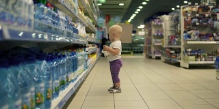 可爱的小孩走在超市的水部，在大市场里挑选瓶子，仔细考虑要买的产品