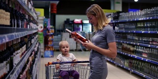 一位年轻貌美的母亲正在超市的饮料部挑选一瓶葡萄酒，而她的小宝宝正坐在购物车里。家庭购物时间