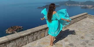 美丽的黑发微笑女孩与健康的长发在吹连衣裙。快乐的年轻女人享受自然，在大海和蓝天上玩。Sveti Stefan、黑山
