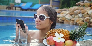 年轻美丽的女人用手机在游泳池旁边的水和盘子与热带水果在慢动作。hd 1920 x1080