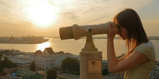 年轻女子透过旅游望远镜，在夕阳下探索城市