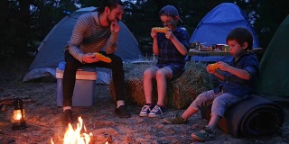 爸爸和儿子吃新鲜的黄色玉米从火在夏季假期，晚餐在附近的火焰自然