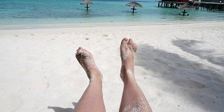 放松的脚与热带岛屿上的白沙嬉戏。POV坐在沙滩座位上