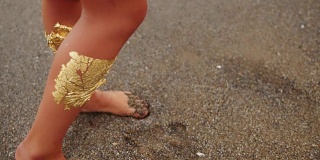 一个性感的女人带着专业的金色妆容看着镜头和观众调情。她摸了摸自己的头发。镜头转向她站在海边潮湿沙滩上的双腿。Slowmotion拍摄