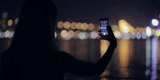 一名女子站在河边用智能手机拍摄夜景