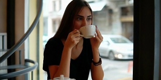 一个美丽的年轻女子，在咖啡店的窗口边喝着热咖啡，梦想着。在漫长忙碌的一天之后休息一下。喝杯热茶放松一下