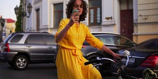 美丽微笑的女人穿着黄色长裙骑着一辆城市自行车，里面有一个篮子和鲜花，用她的手机拍摄。替身。Slowmotion